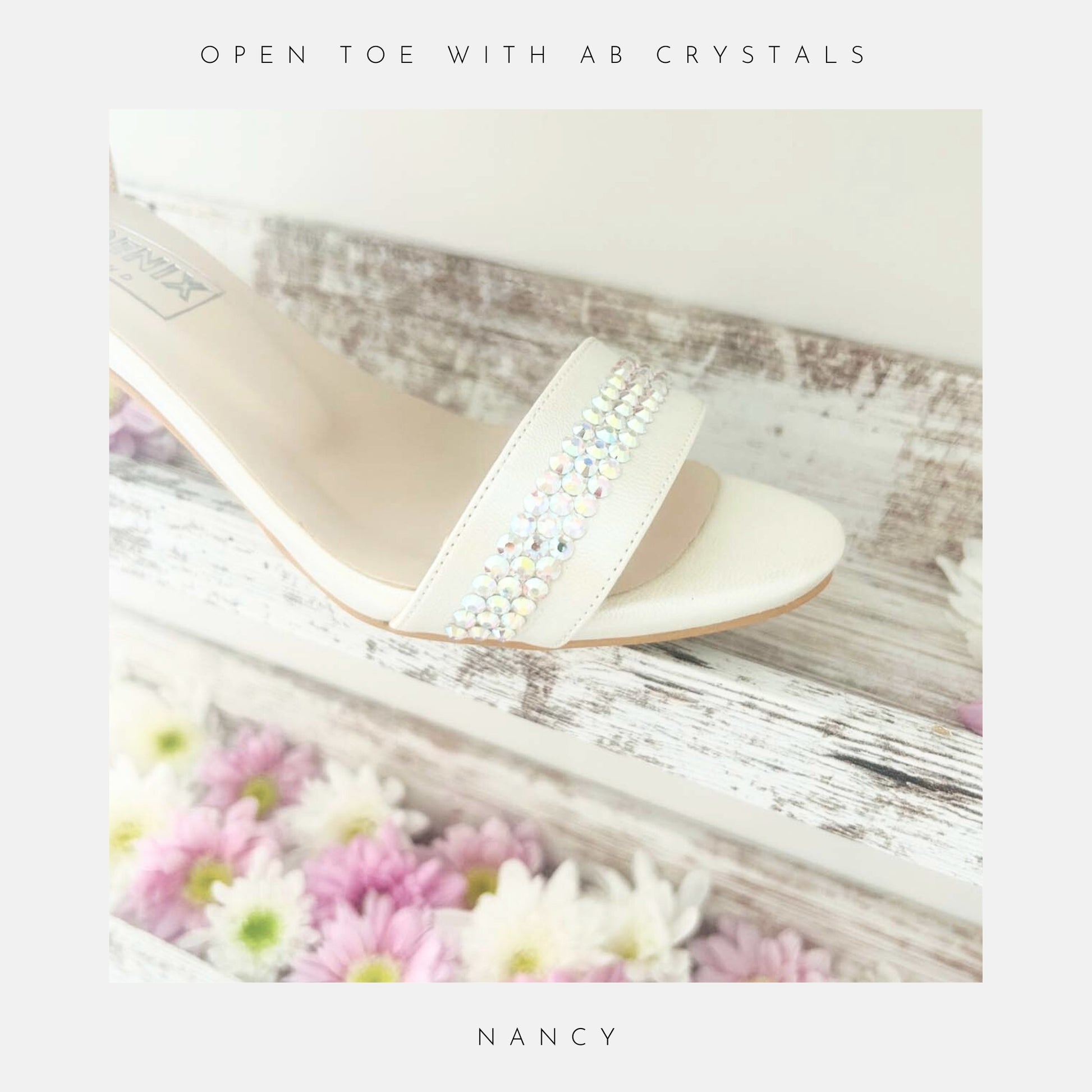 An ivory sandal, the Nancy Crystal Kitten Heel Bridal Shoes by Phoenix England, features pearl vegan leather and a crystal embellished strap. Displayed on a rustic white shelf with pastel daisies. “HAND FINISHED WITH CRYSTALS” above and “NANCY” below. Perfect for weddings.