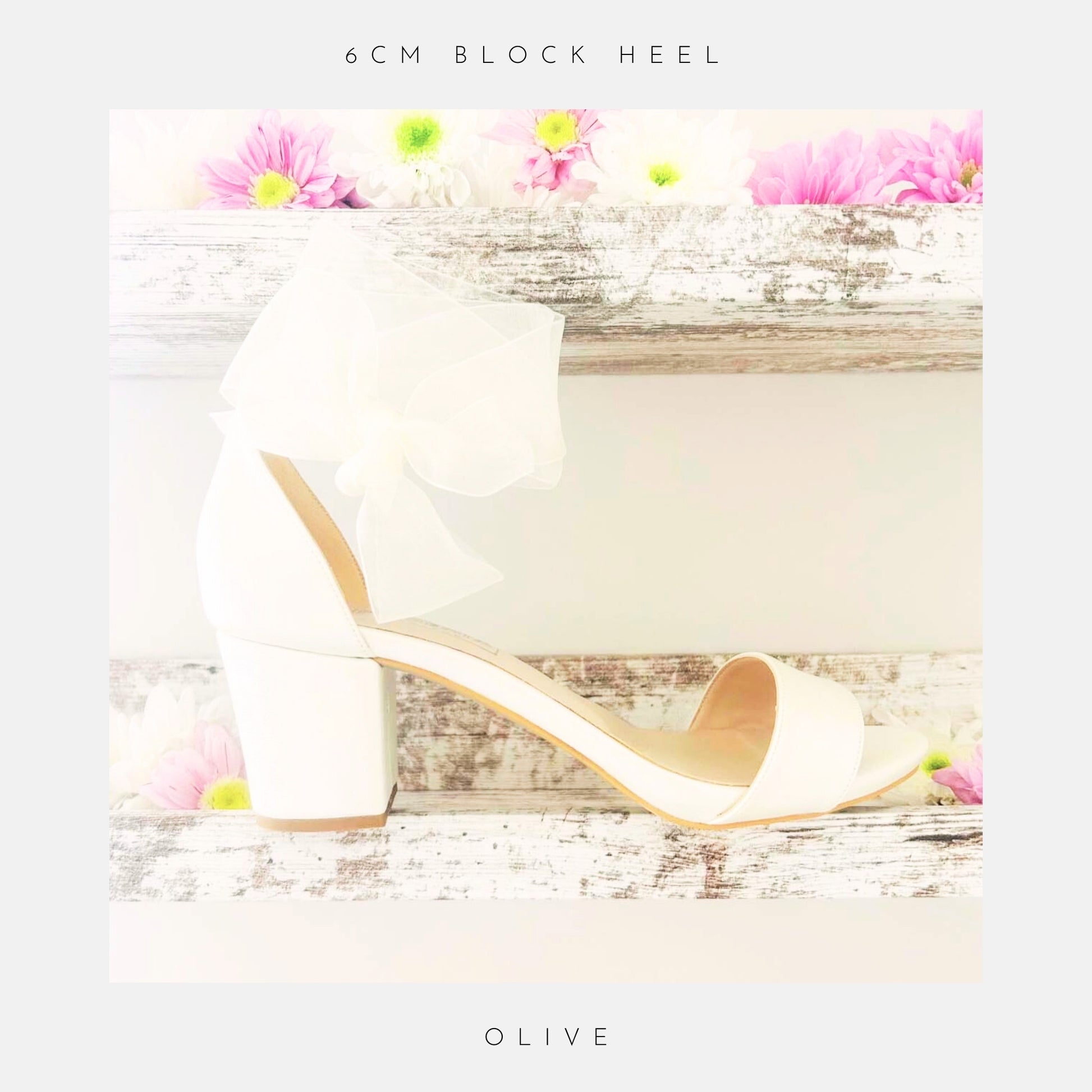 The Phoenix England Olive Block Heel Bridal Shoes Organza Strap, an ivory block heel bridal shoe with an organza bow, is displayed on a rustic white wooden shelf with pink and white flowers. Text: 6cm block heel & Olive.