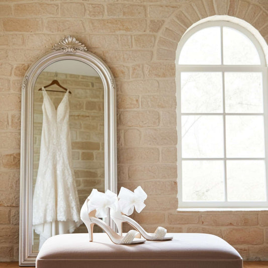 Wedding dress hanging in a mirror with ivory high-heeled shoes on a bench in a room with a brick wall and window.