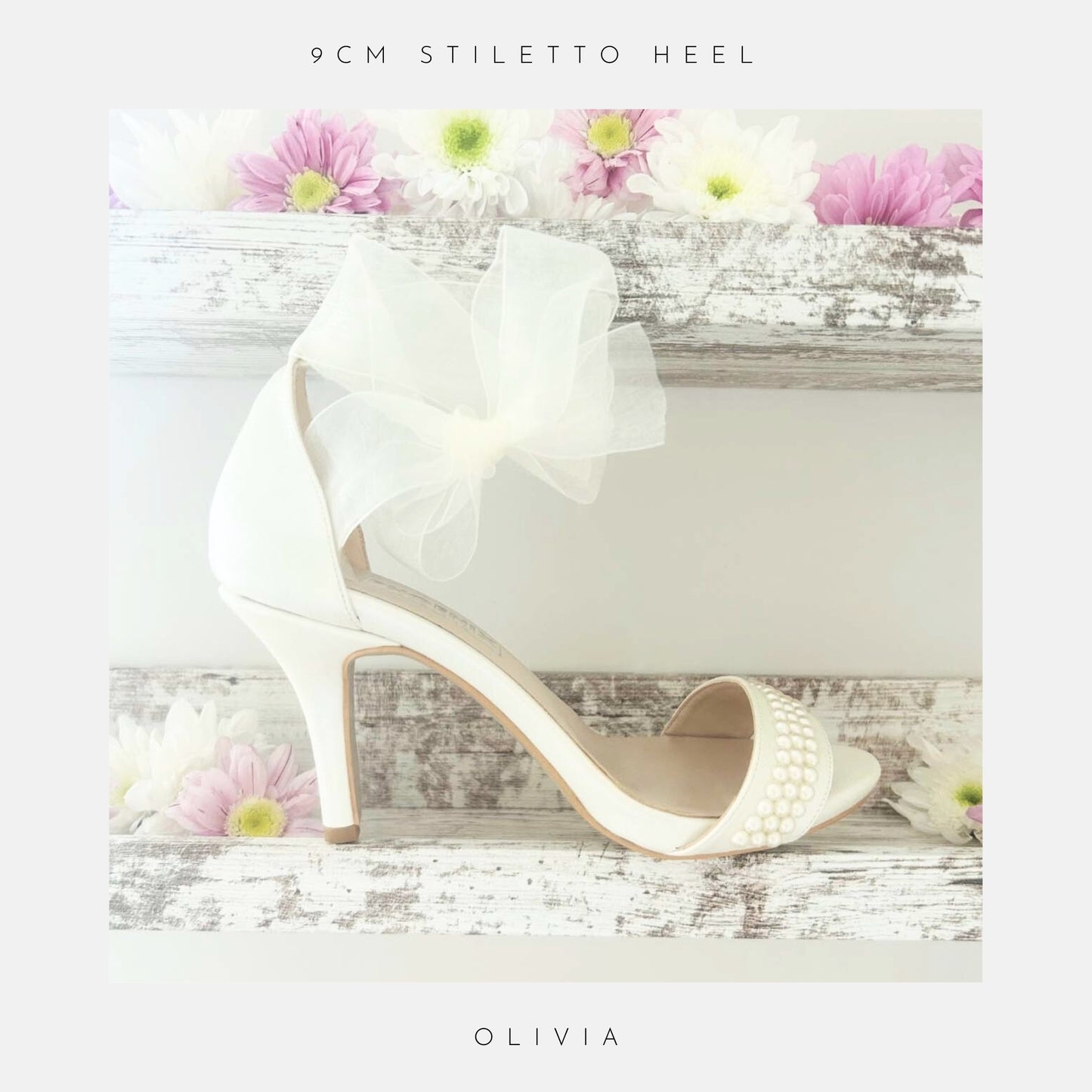 The Olivia Organza Strap Pearl Bridal Shoes by Phoenix England are elegant ivory heels with pearl accents and a large organza bow, perfect for brides, shown on rustic wooden shelves adorned with pink, purple, and white flowers.