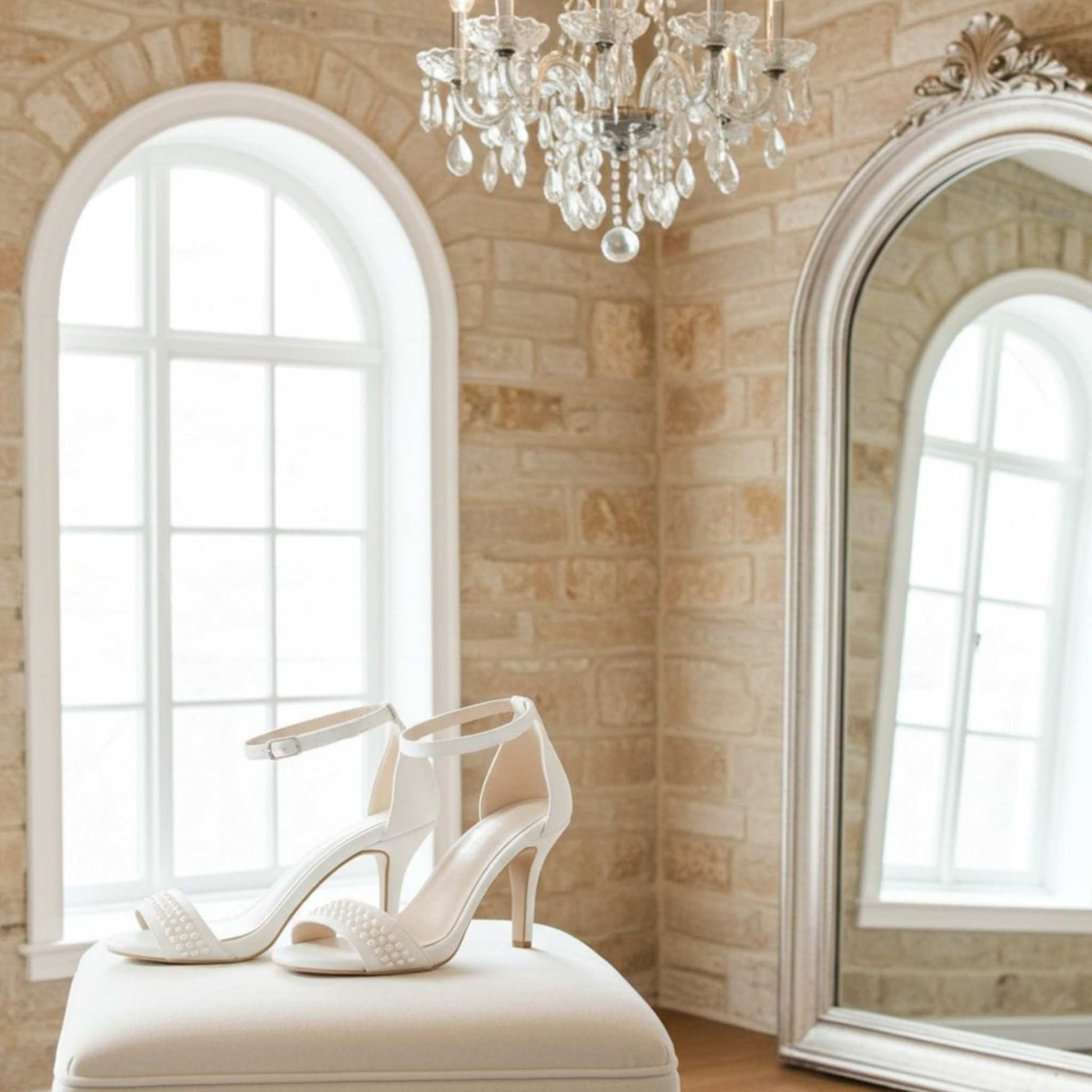 Ivory high-heeled shoes on a white stool in a room with a chandelier and large mirror.
