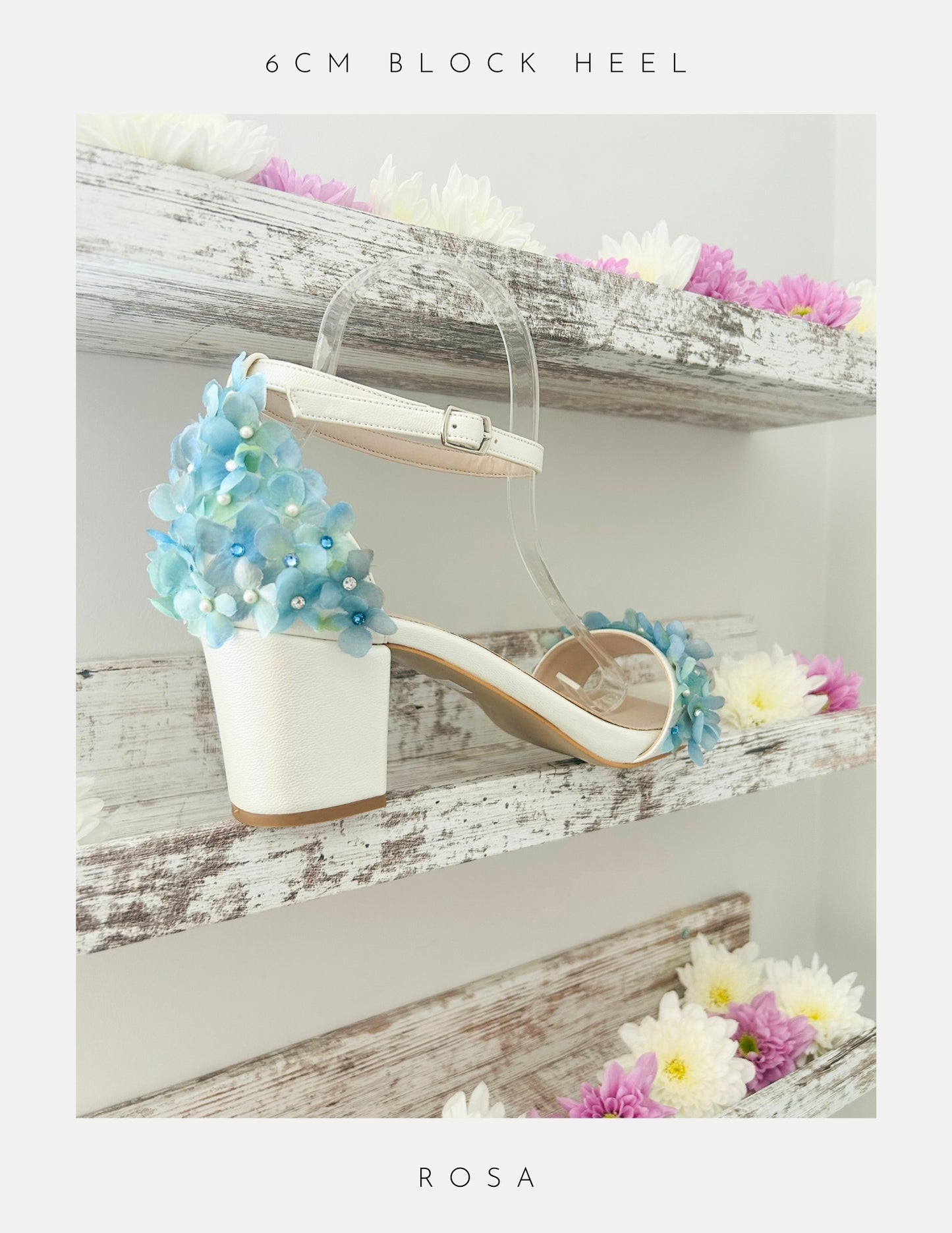 A white 6CM Rosa Floral Low Block Heel Bridal Shoe by Phoenix England with blue and white floral heel embellishments sits on a rustic shelf decorated with colorful daisies—perfect chic bridal shoes.