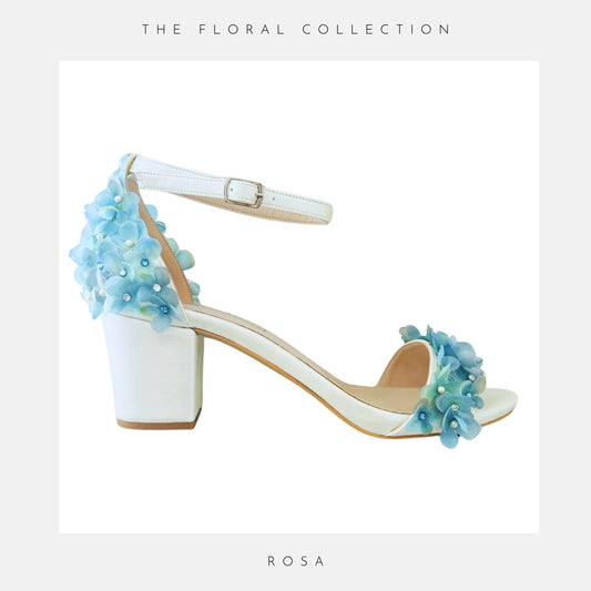 An ivory block heeled sandal with ankle strap, featuring blue floral embellishments on the heel and toe strap. “The Floral Collection” appears at the top.