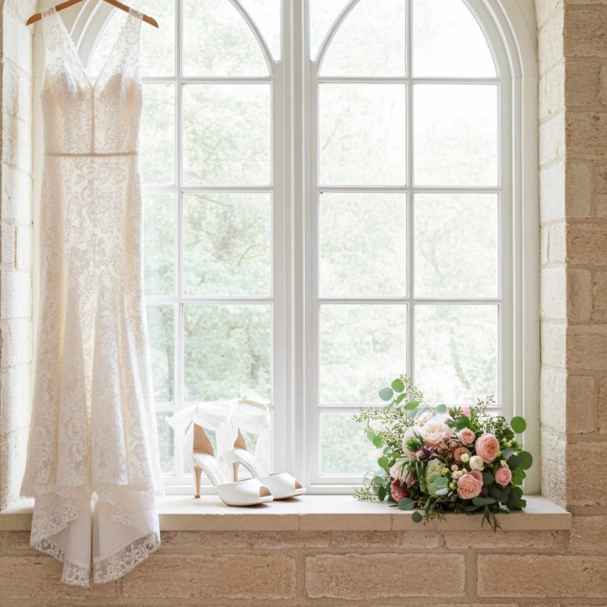 Ivory lace wedding dress on a hanger, ivory peep toe high-heeled bridal shoes, and a bouquet of flowers on a windowsill.