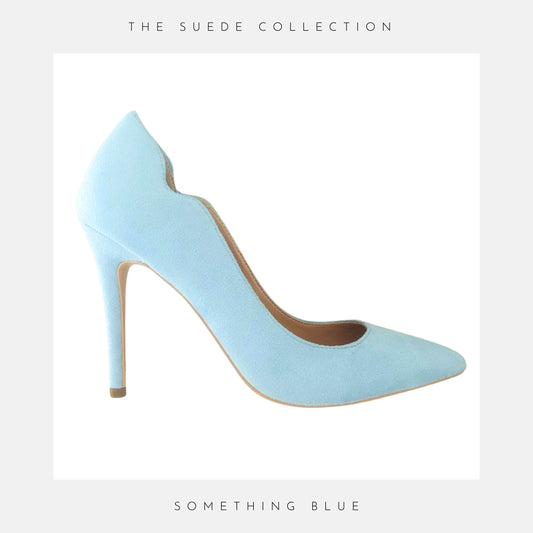 Blue high-heeled bridal shoe on a wooden surface with flowers, labelled 'The Suede Collection'.