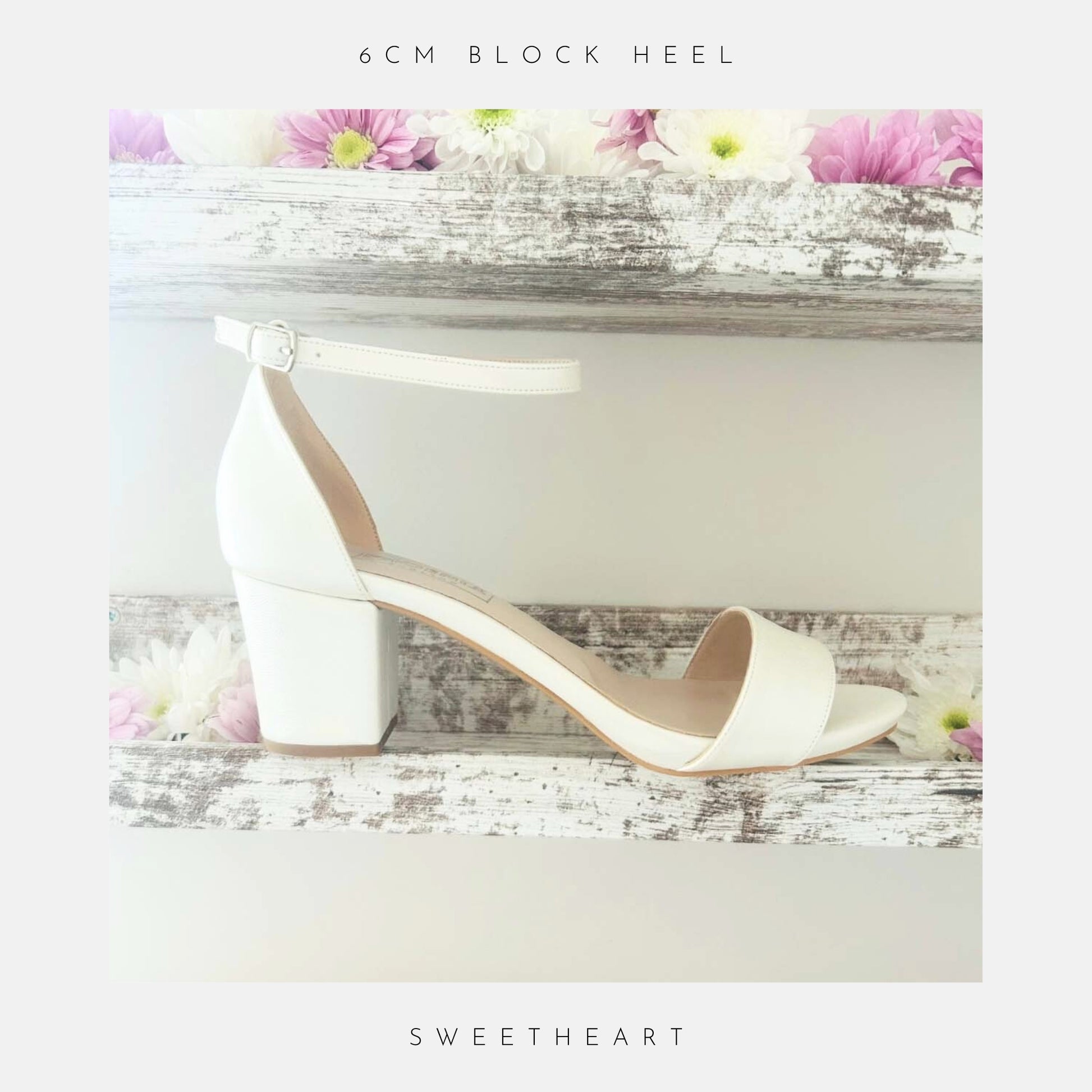 The Phoenix England Sweetheart Plain Low Block Heel Bridal Shoes, in white vegan leather with an ankle strap and open toe, are displayed on a rustic wooden shelf with pink and white flowers. "6cm block heel" is above, "Sweetheart" below.