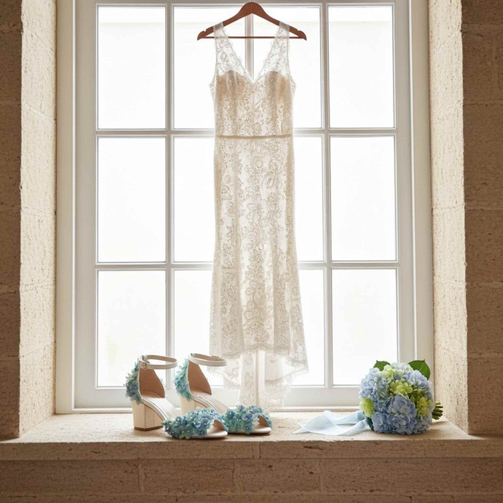 Wedding dress hanging by a window with peep toe floral shoes and bouquet on a windowsill.