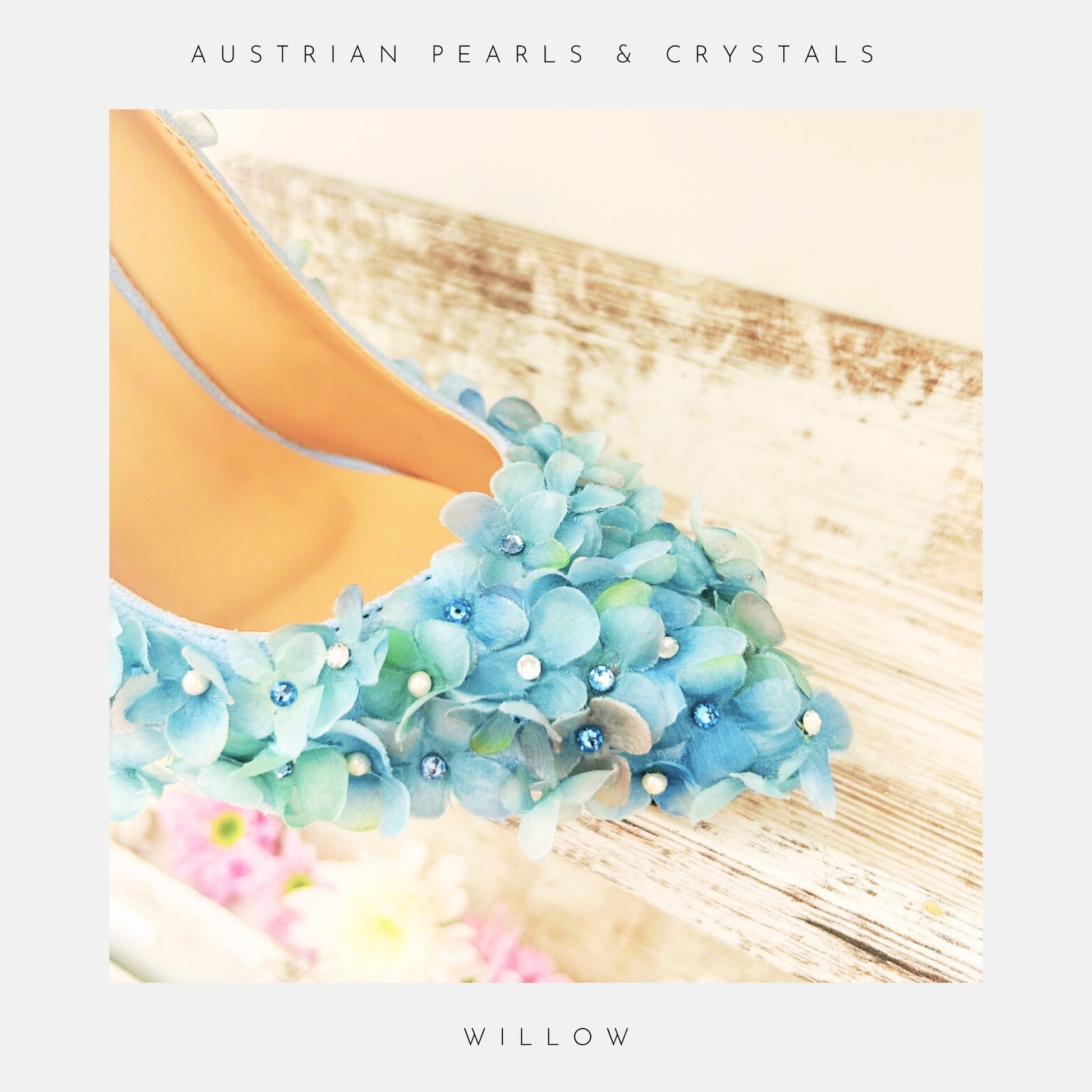 Close-up of Willow Pastel Blue Floral Bridal Shoes by Phoenix England, adorned with blue-green fabric flowers, pearls, and crystals on a rustic wooden surface. Text reads Phoenix England above and Willow below—a stunning piece of floral bridal footwear.