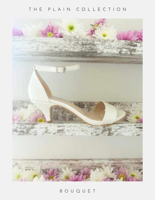 A pair of Bouquet Kitten Heel Bridal Shoes by Phoenix England in pearl vegan leather with an ankle strap rests on a rustic white shelf, surrounded by pink and white daisies. The Plain Collection—perfect bridal heels for your special day.