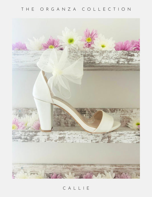 Ivory high-heeled shoe with a bow on a wooden surface with flowers, labeled 'The Organza Collection' and 'Callie'.