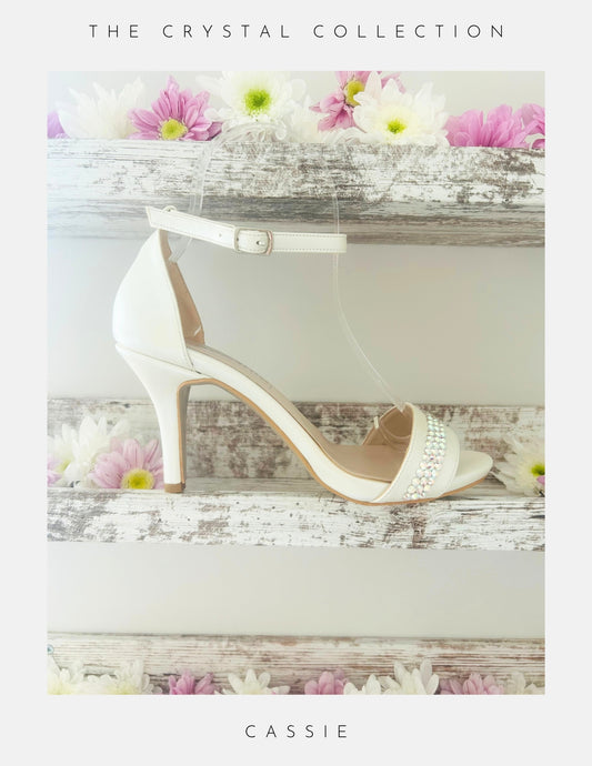 Ivory bridal sandal with crystals on a wooden surface with flowers, labelled 'Cassie' from 'The Crystal Collection'.