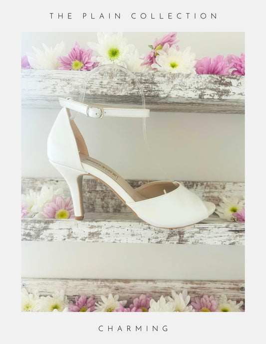 Ivory high-heeled peep toe bridal shoe on a wooden surface with flowers, labelled 'The Plain Collection'.