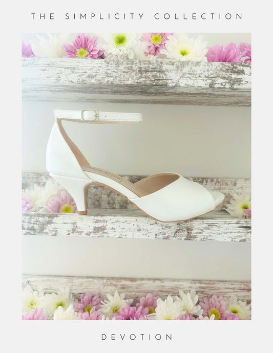 The Devotion Peep Toe Kitten Heel Bridal Shoes by Phoenix England sit on a rustic white shelf, surrounded by pastel pink and white flowers. Text reads, “The Simplicity Collection – Devotion.”.