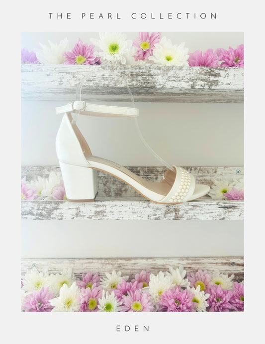 The Eden Pearl Low Block Heel Bridal Shoes by Phoenix England feature elegant pearl accents and an ivory block heel, displayed on a rustic shelf with pink and white flowers. Part of The Pearl Collection, perfect for brides. Text below reads Eden.