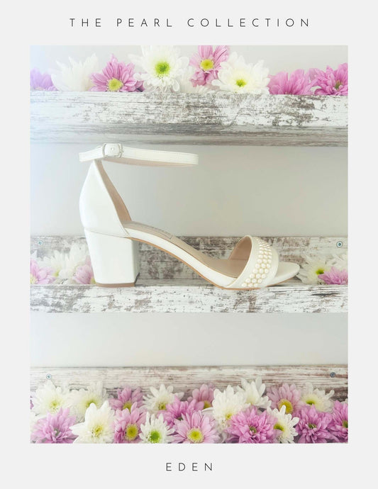 The Eden Pearl Low Block Heel Bridal Shoes by Phoenix England feature elegant pearl accents and an ivory block heel, displayed on a rustic shelf with pink and white flowers. Part of The Pearl Collection, perfect for brides. Text below reads Eden.