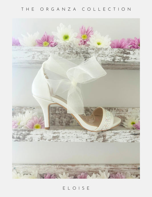 Ivory high-heeled wedding shoes with a bow on a wooden surface with flowers, labelled 'The Organza Collection' and 'Eloise'.