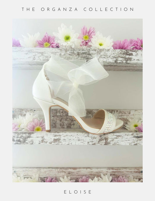 Ivory high-heeled wedding shoes with a bow on a wooden surface with flowers, labelled 'The Organza Collection' and 'Eloise'.
