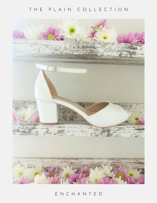 Ivory mid heel block peep toe bridal shoe on a wooden surface with flowers, labelled 'The Plain Collection' and 'Enchanted'.