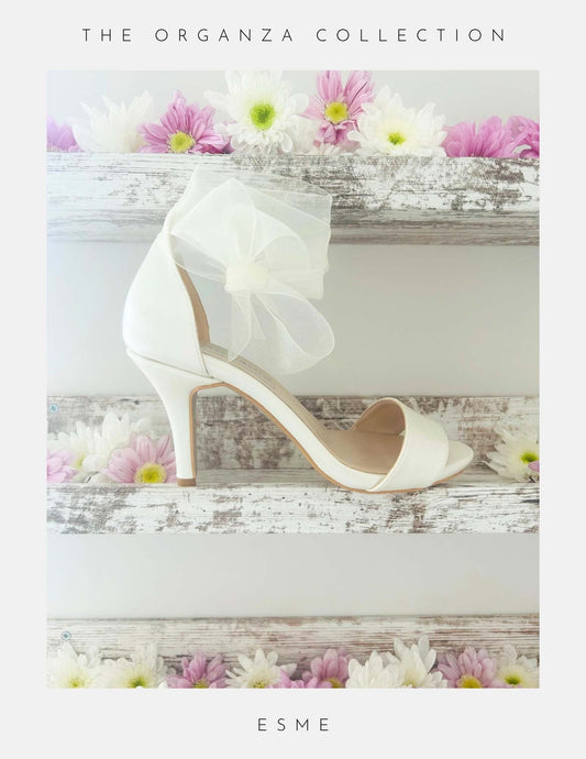Ivory high-heeled bridal open toe shoe with a bow on a wooden surface with flowers, labelled 'The Organza Collection' and 'Esme'.