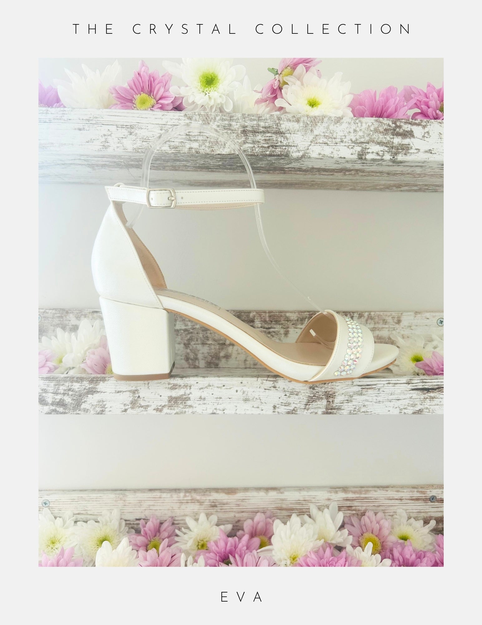 Displayed on a rustic white wooden shelf with pink and white flowers, the Phoenix England Eva Crystal Low Block Heel Bridal Shoes in vegan leather feature a white ankle strap and crystal accents. Text above: The Crystal Collection; below: Eva.