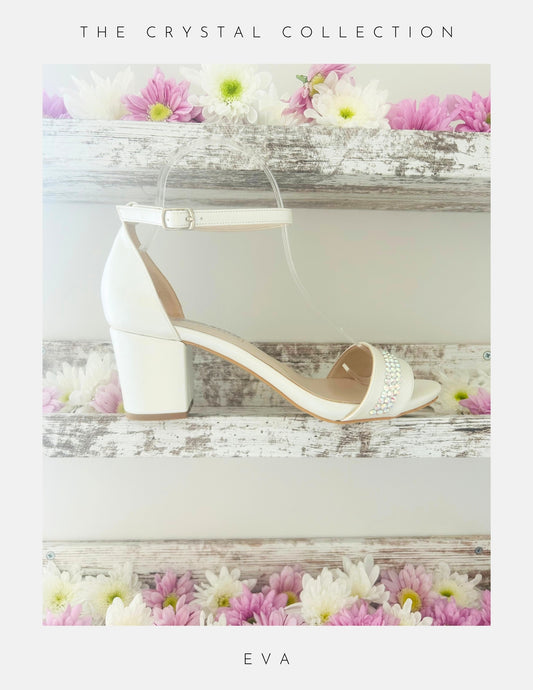 Displayed on a rustic white wooden shelf with pink and white flowers, the Phoenix England Eva Crystal Low Block Heel Bridal Shoes in vegan leather feature a white ankle strap and crystal accents. Text above: The Crystal Collection; below: Eva.
