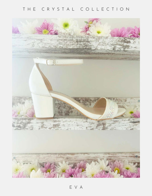 Displayed on a rustic white wooden shelf with pink and white flowers, the Phoenix England Eva Crystal Low Block Heel Bridal Shoes in vegan leather feature a white ankle strap and crystal accents. Text above: The Crystal Collection; below: Eva.