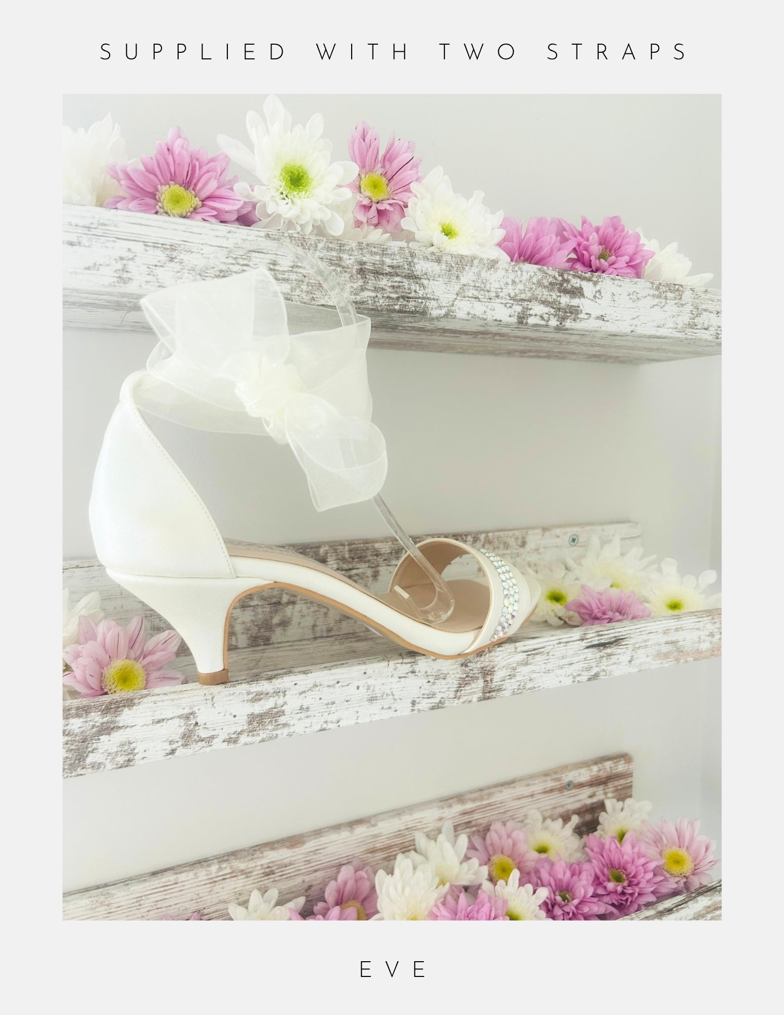 The Phoenix England Eve Organza Kitten Heel Bridal Shoes, featuring a sheer bow and organza strap, are displayed on a rustic white shelf with pink and white flowers. Text above reads Supplied with Two Straps and below, Eve.