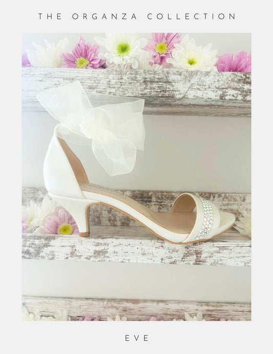 An ivory Phoenix England Eve Organza Kitten Heel Bridal Shoe with a sheer bow and sparkling rhinestones sits on rustic wooden shelves adorned with pink and white daisies. “The Organza Collection” appears above and “Eve” below.