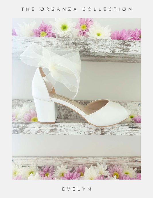 Ivory peep toe wedding shoes with a large bow and mid block heel on a floral background, featuring 'The Organza Collection' and 'Evelyn'.