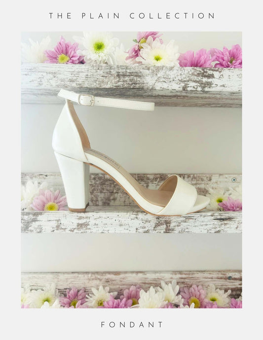 Ivory tall block high-heeled shoe on a wooden surface with flowers in the background, labelled 'The Plain Collection' and 'Fondant'.