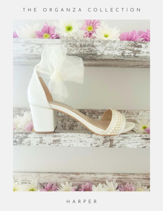 Ivory bridal sandal with pearl details on a wooden surface with flowers, labelled 'The Organza Collection' and 'Harper'.