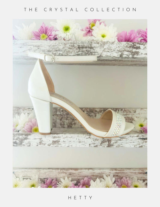 Ivory high-heeled block heel wedding shoe with crystal embellishments on a decorative background with flowers.