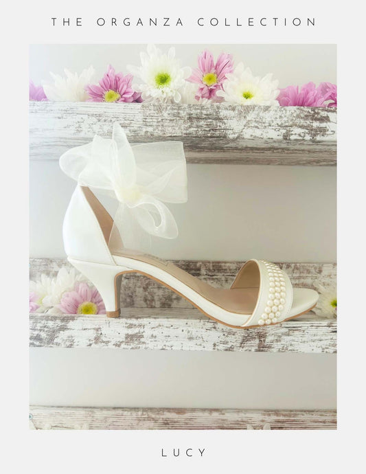 Ivory open toe pearl bridal sandal with pearls on a wooden surface with flowers, labelled 'The Organza Collection' and 'Lucy'.