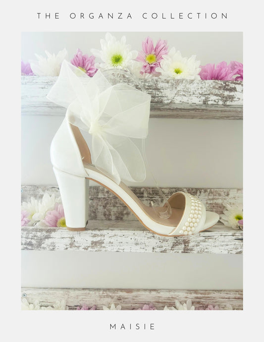 Ivory high-heeled bridal shoe with a decorative bow and pearl details on a wooden surface with flowers.