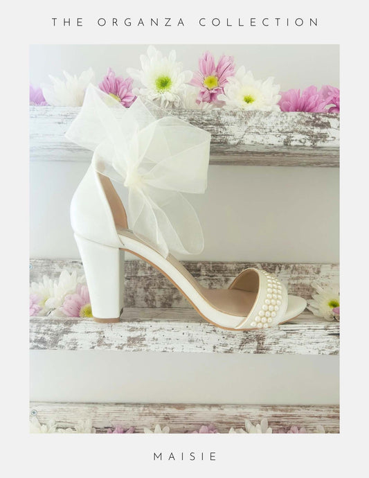 Ivory high-heeled bridal shoe with a decorative bow and pearl details on a wooden surface with flowers.