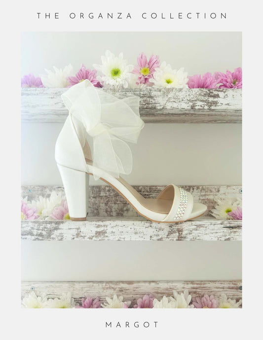 Ivory bridal sandal with an organza bow and crystals to the front on a wooden surface with flowers