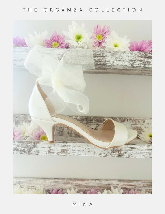 Ivory open toe kitten heel bridal sandal with an organza bow on a wooden surface with flowers, labelled 'The Organza Collection' and 'Mina'.