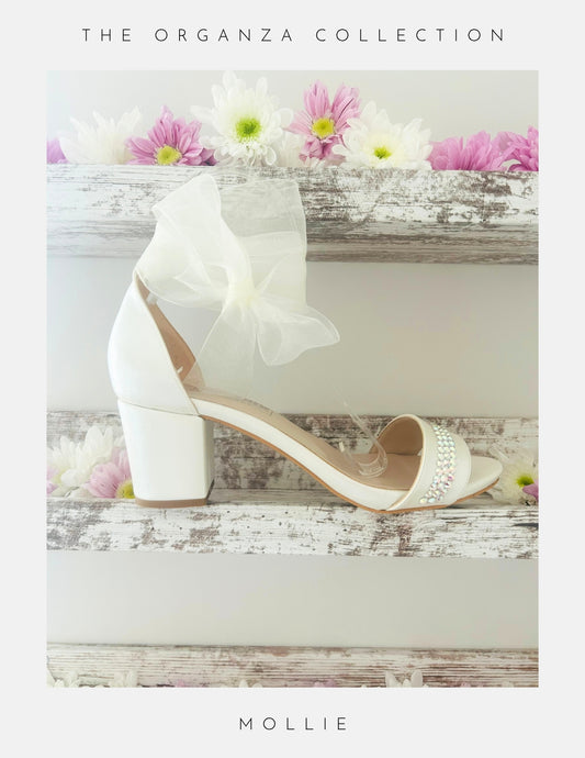 Ivory bridal sandal with block heel organza bow and crystals on a wooden surface with flowers, labelled 'The Organza Collection' and 'Mollie'.