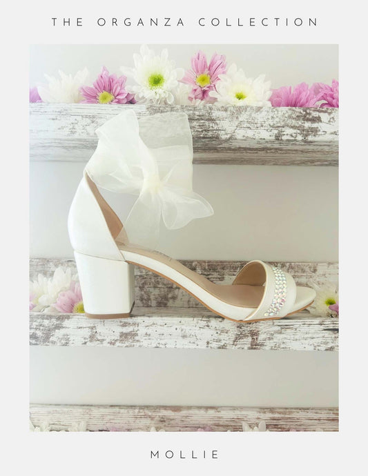 Ivory bridal sandal with block heel organza bow and crystals on a wooden surface with flowers, labelled 'The Organza Collection' and 'Mollie'.