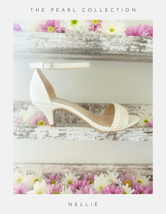 An ivory Nellie Pearl Kitten Heel Bridal Shoe by Phoenix England with an ankle strap and pearl embellishments sits on rustic wooden shelves adorned with pink and white daisies. Text above: The Pearl Collection; below: Elegant bridal shoes.