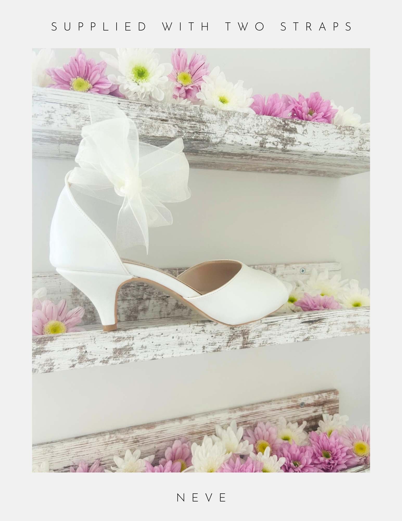 Ivory wedding shoe with a bow and kitten heel on a wooden surface with flowers, labelled 'Supplied with two straps' and 'Neve'.