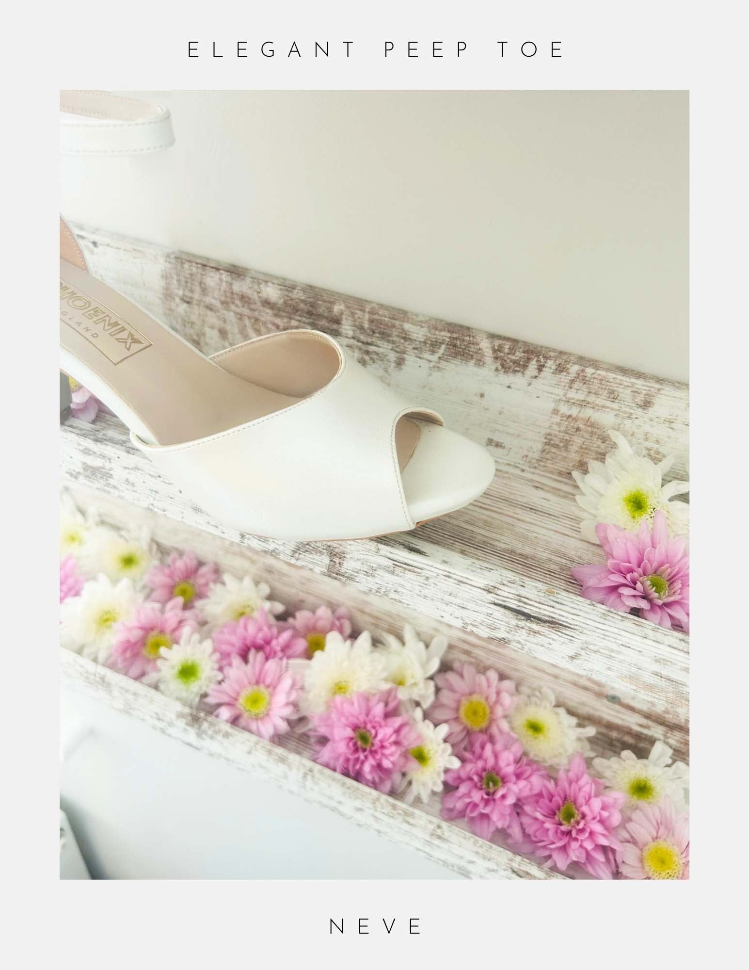 Close up of peep toe ivory kitten heel bridal shoe on a wooden surface with flowers, labelled 'Elegant Peep Toe' and 'Neve'.
