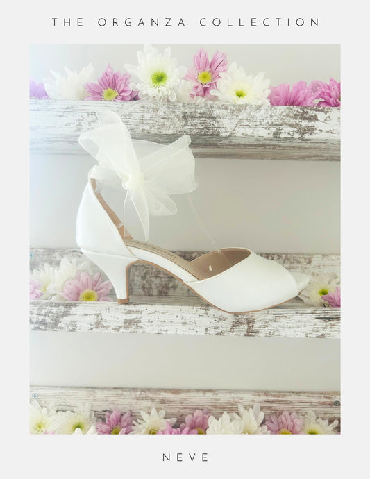 The Neve Organza Strap Kitten Heel Bridal Shoes by Phoenix England are displayed on a rustic white shelf with pink, white, and yellow flowers. Text reads: The Organza Collection and Neve.