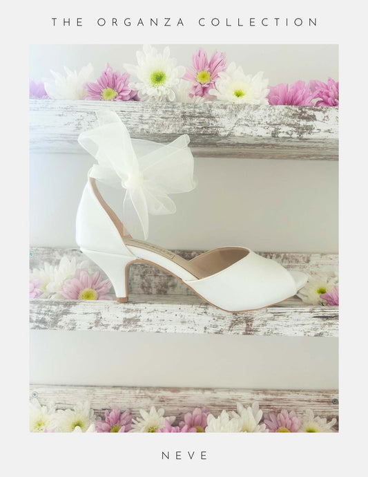 Ivory peep toe wedding shoe with an organza bow and kitten heel on a wooden surface with flowers, labelled 'The Organza Collection'.