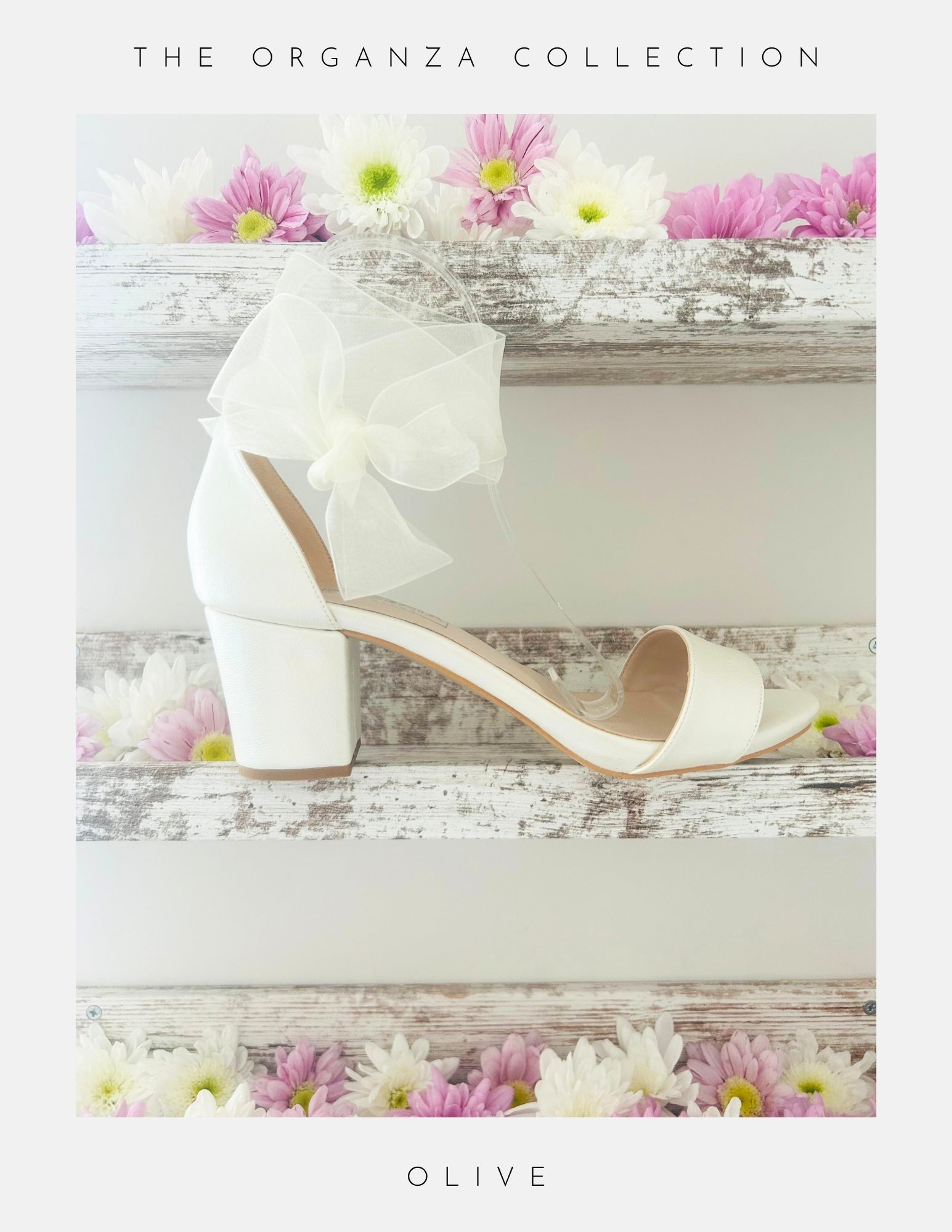 The Phoenix England Olive Block Heel Bridal Shoes Organza Strap, an ivory block heel bridal shoe with an organza bow, is displayed on a rustic white wooden shelf with pink and white flowers. Text: The Organza Collection.