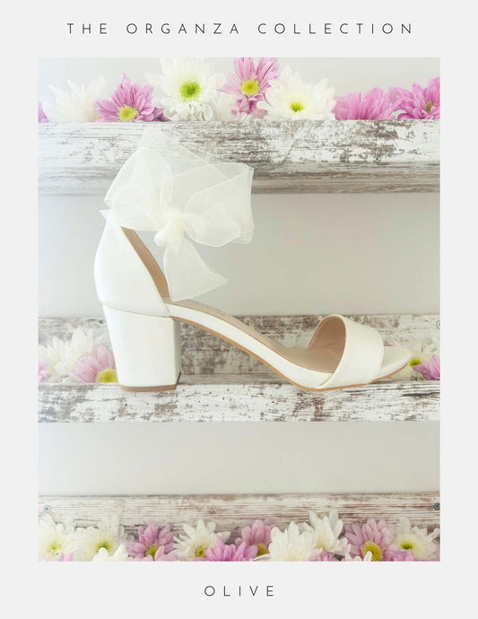 The Phoenix England Olive Block Heel Bridal Shoes Organza Strap, an ivory block heel bridal shoe with an organza bow, is displayed on a rustic white wooden shelf with pink and white flowers. Text: The Organza Collection.