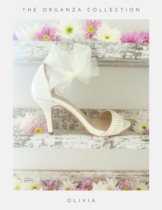 The Olivia Organza Strap Pearl Bridal Shoes by Phoenix England are elegant ivory heels with pearl accents and a large organza bow, perfect for brides, shown on rustic wooden shelves adorned with pink, purple, and white daisies. The Organza Collection.