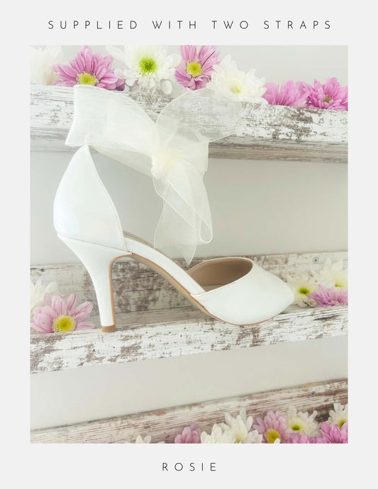 Ivory high-heeled shoe with a decorative bow on a wooden surface with flowers.