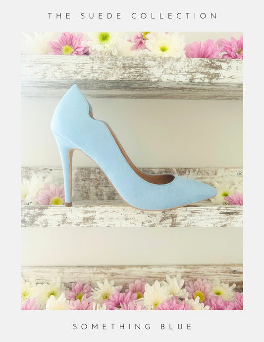Blue high-heeled bridal shoe on a wooden surface with flowers, labelled 'The Suede Collection'.