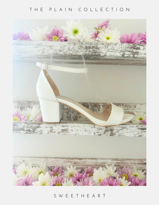 Ivory wedding sandal with block heel on a wooden surface with flowers, labelled 'The Plain Collection' and 'Sweetheart'.